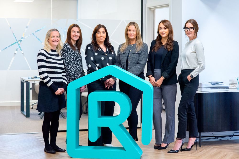 A group gathers around a large turquoise letter R logo display in a modern office setting.