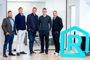 A group of five professionals stand together in a modern office space next to a large turquoise company logo.