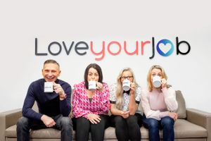 A group of four coworkers sitting on a couch drinking coffee under a Love Your Job sign on the wall. Corporate photoshoot