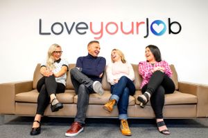 A group of coworkers sitting on a beige couch laughing together under a Love Your Job sign on the wall.