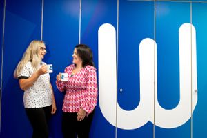 Two people chat and laugh while standing against a bright blue wall with a large white letter W. Corporate photoshoot office shots