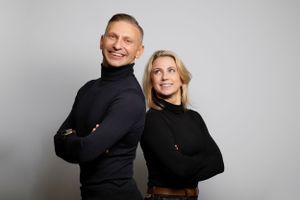 Two people wearing black turtleneck sweaters pose together against a gray background.