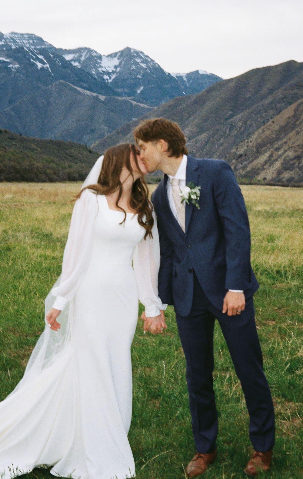 Bride and groom kissing in a green meadow with snow-capped mountains backdrop on their wedding day.