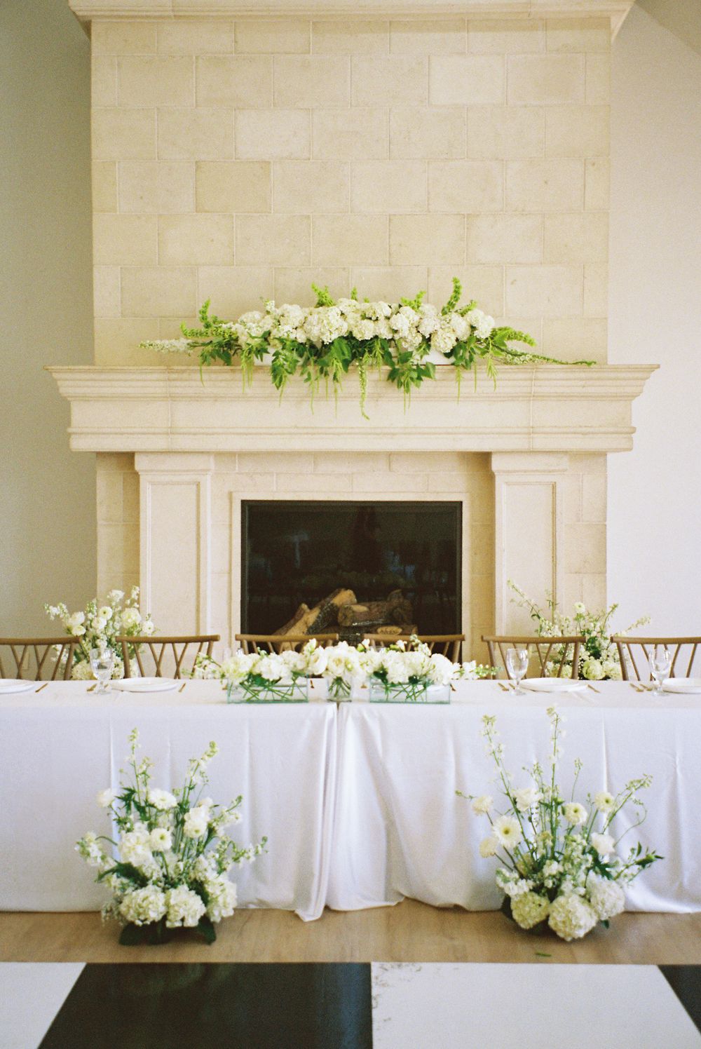 Elegant wedding reception head table with white floral arrangements before a stone fireplace.