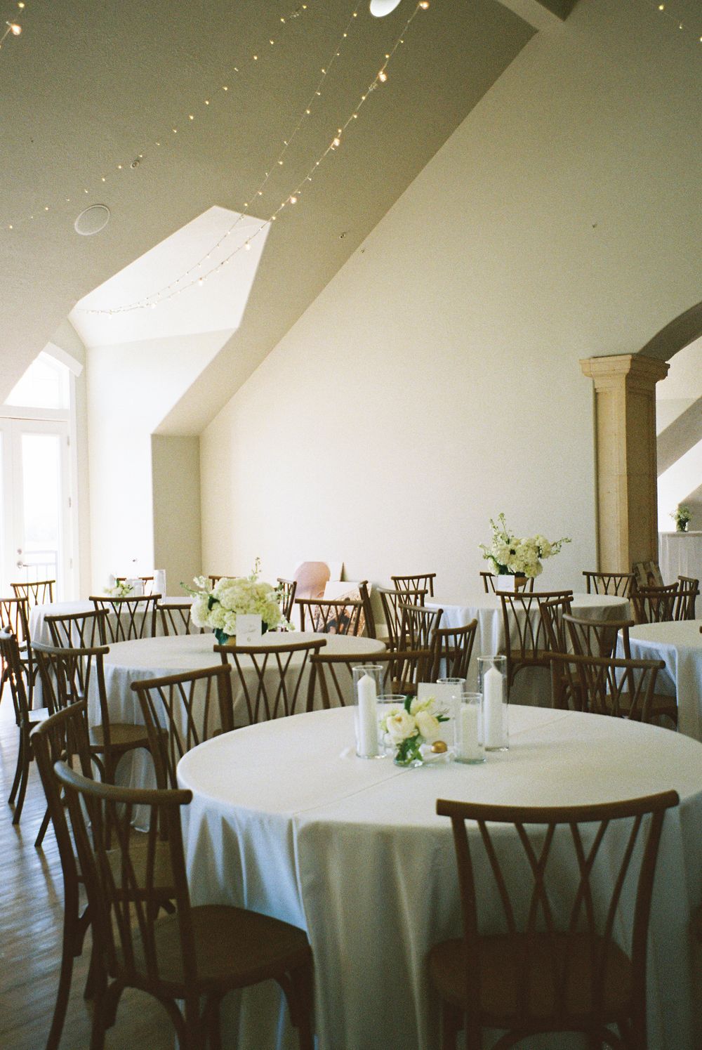 Bright wedding reception hall with round tables, wooden chairs, white linens, and string lights.