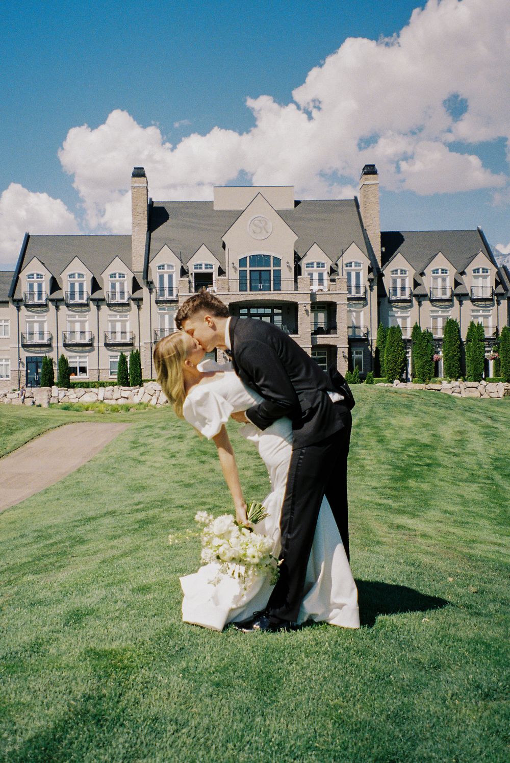 Groom dipping bride for a kiss on green lawn before a grand estate wedding venue.