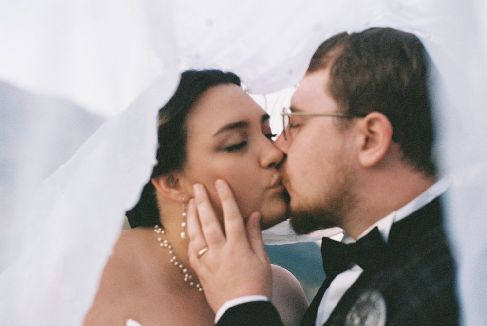 Bride and groom share a tender kiss under flowing white veil, bride wearing pearl necklace.