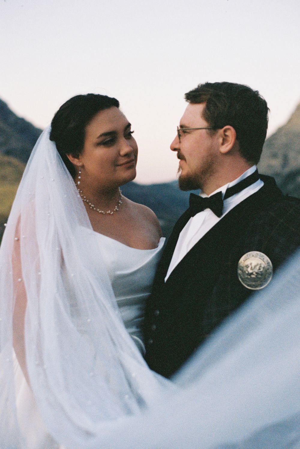 Bride in white veil and groom in tuxedo share an intimate gaze with mountains in background.