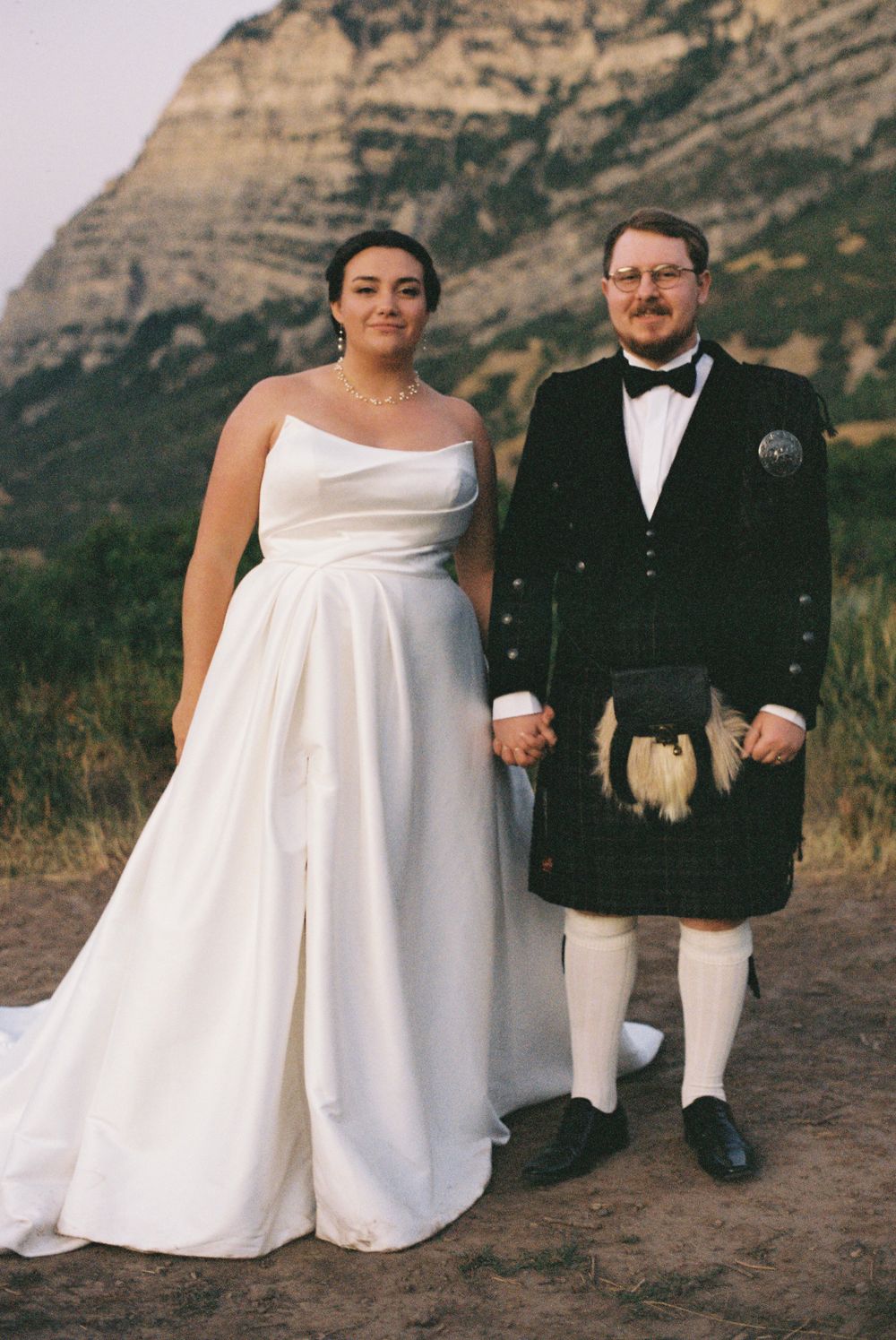 Bride and groom hold hands posing before dramatic rocky mountain cliff in wedding attire.