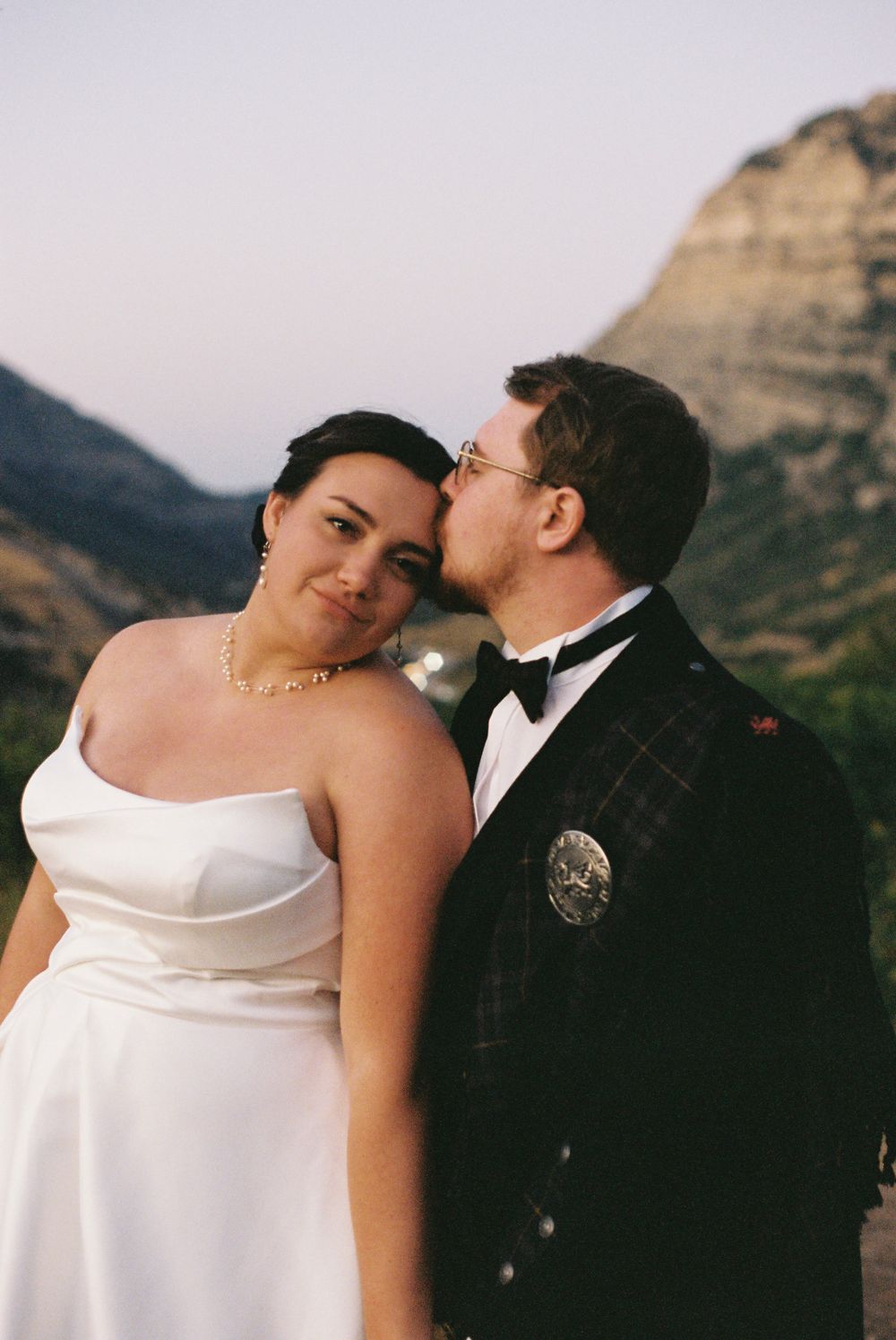 Groom in Scottish kilt kisses bride on cheek as she smiles with rocky mountain backdrop at dusk.