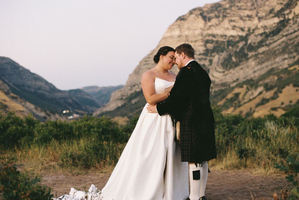 Newlyweds touch foreheads tenderly in a romantic embrace with majestic mountain canyon behind them.