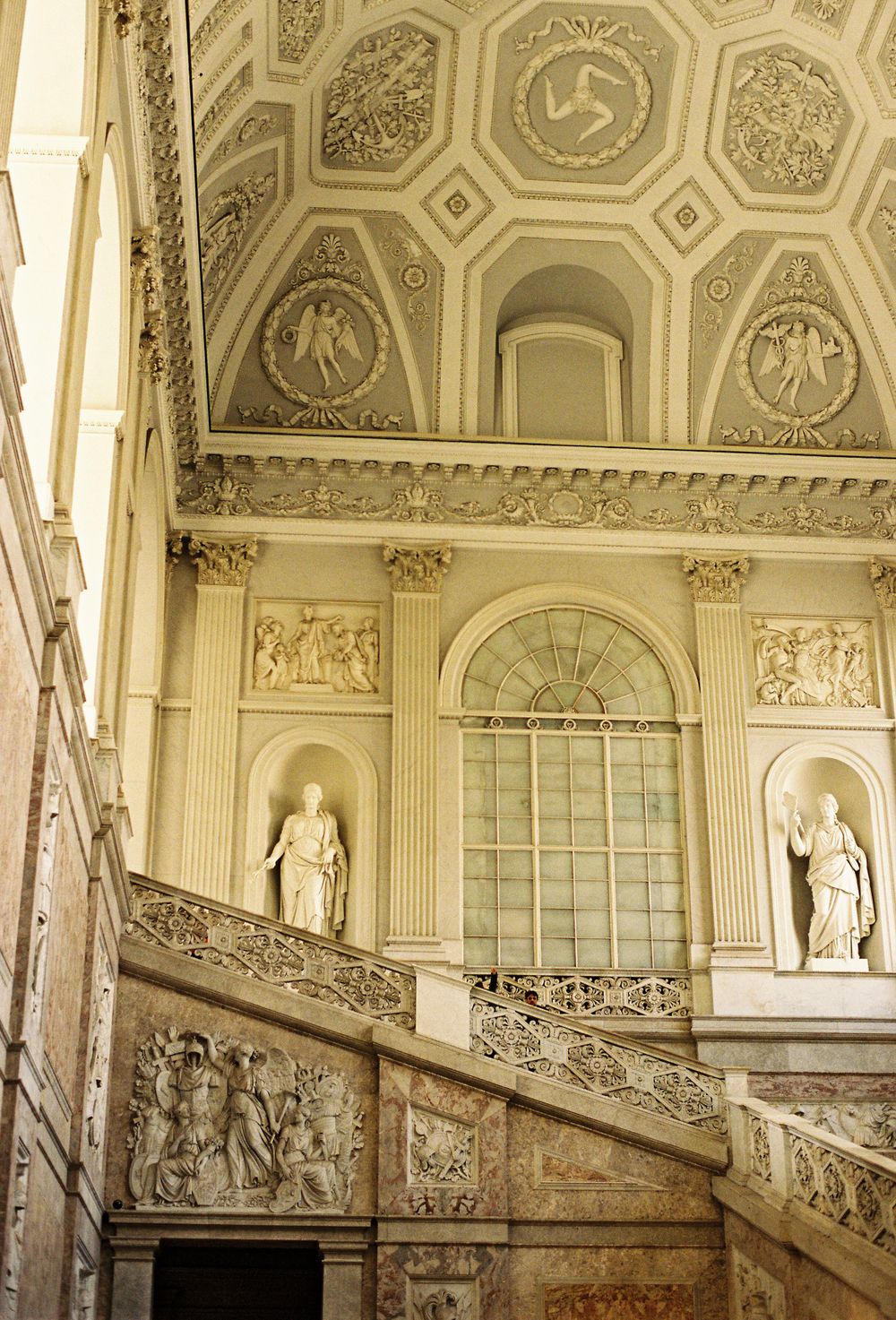 Ornate baroque staircase with sculpted reliefs, classical statues, and decorated coffered ceiling in a grand palace.
