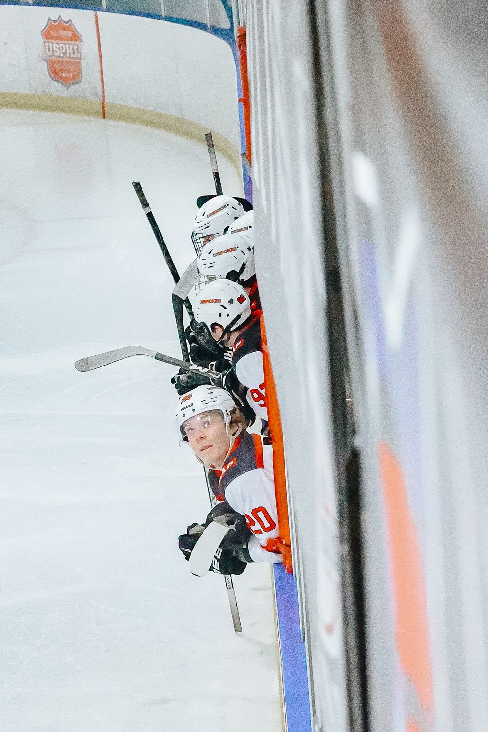 Hockey players lean against the boards during a game, with white jerseys and helmets visible against the arena background.