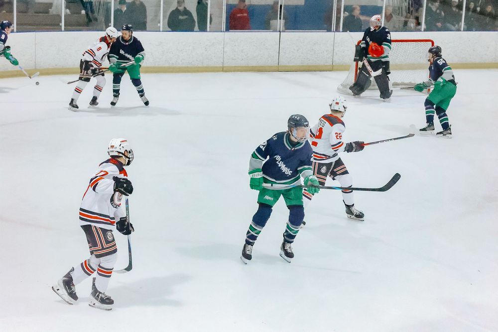Hockey players in blue and green jerseys compete for the puck on an indoor ice rink during a game.