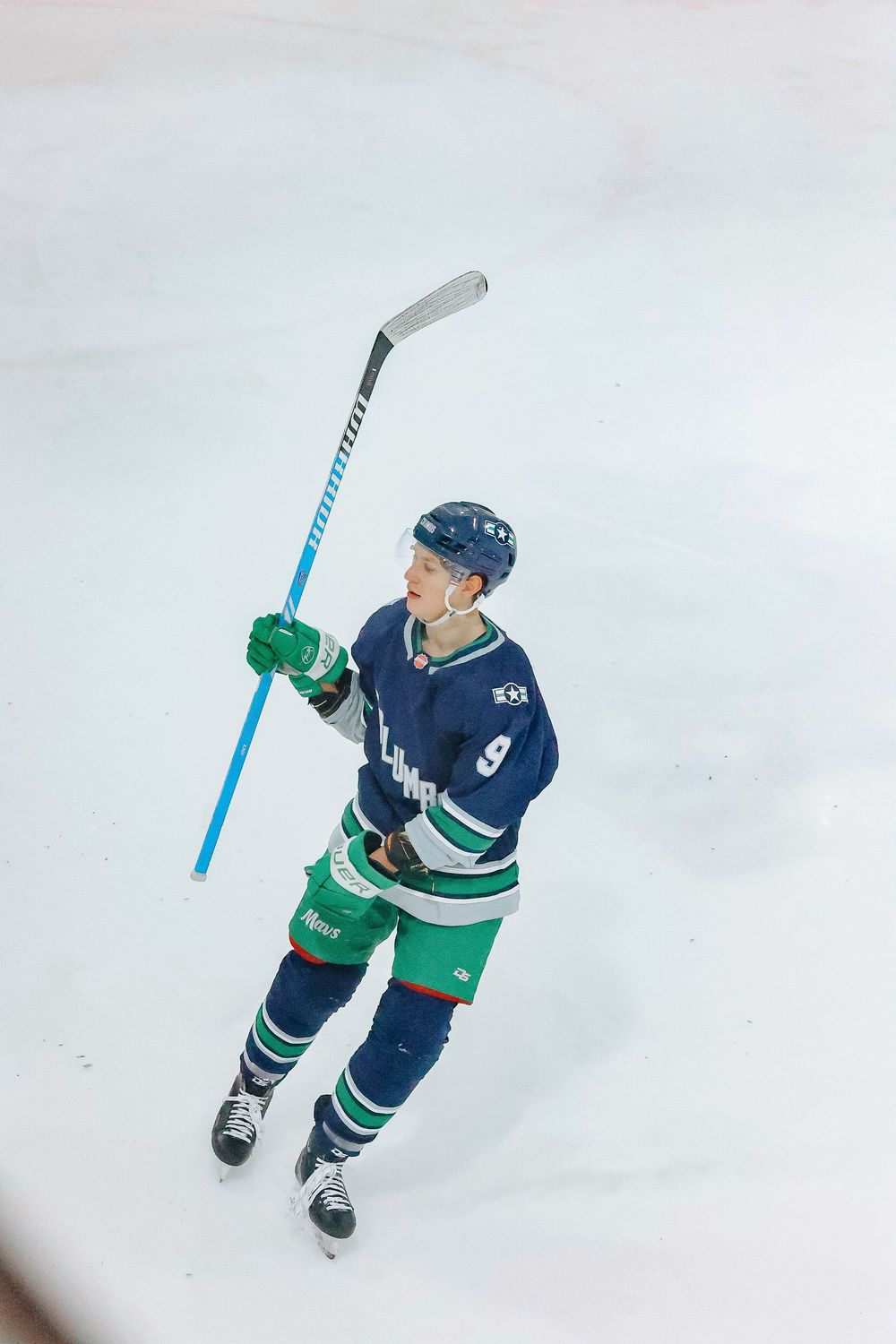 Hockey player in navy and green uniform raises stick during game on ice rink.