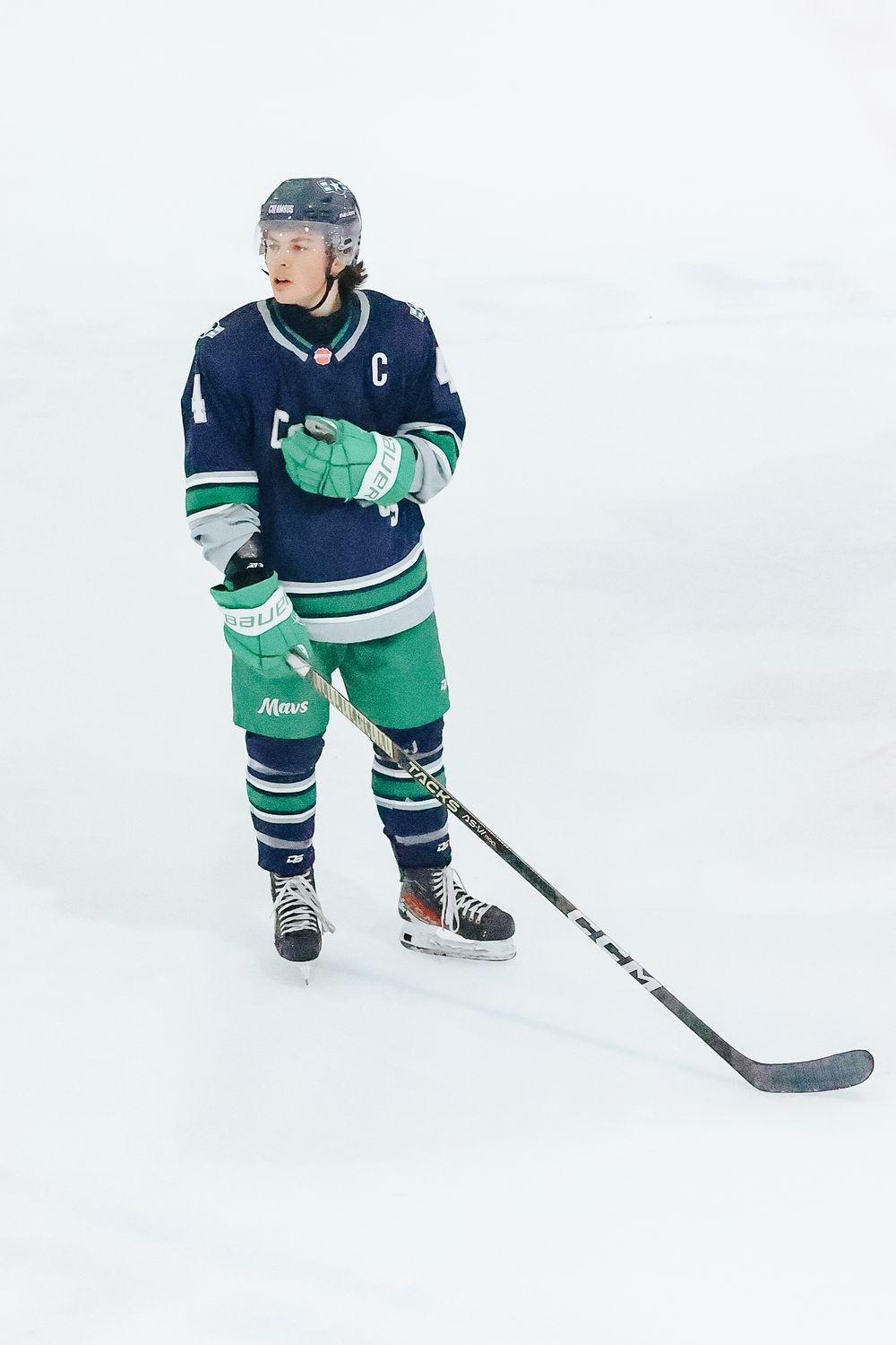 Hockey player in navy blue and green jersey skates on ice while holding a hockey stick.