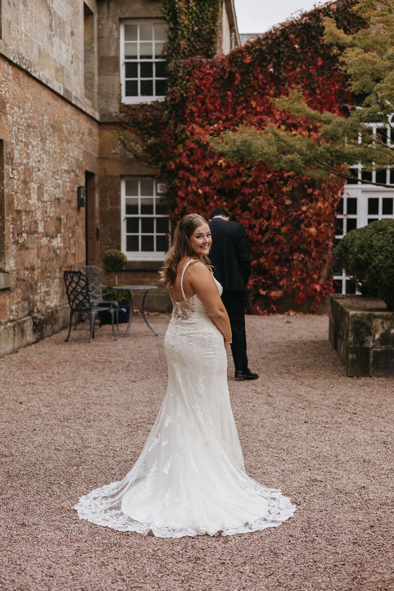 A bride in a flowing white lace wedding dress poses in front of a stone building with red autumn vines.