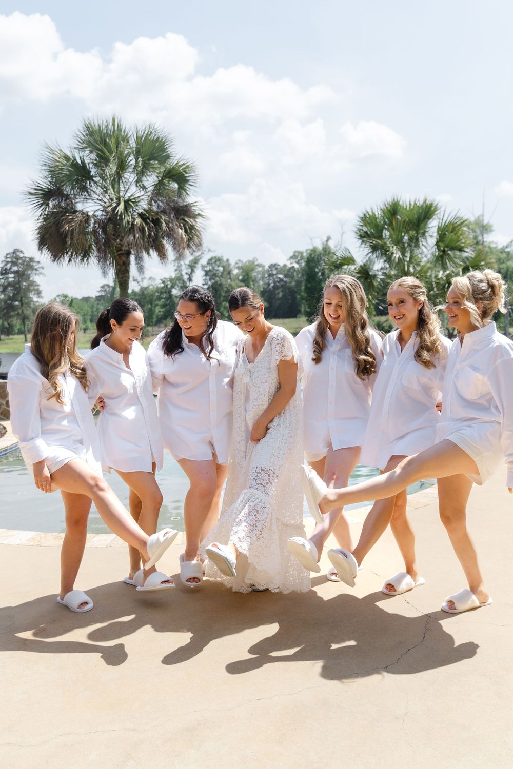 Bride and bridesmaids in white shirts kick up their heels together near a pool with palm trees.