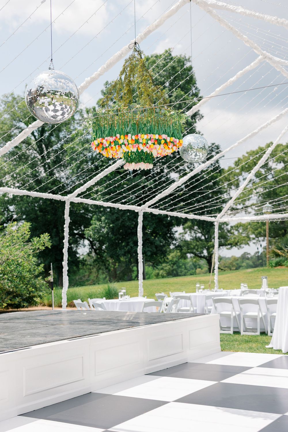 Outdoor wedding reception tent with checkered dance floor, disco balls, and floral chandelier.