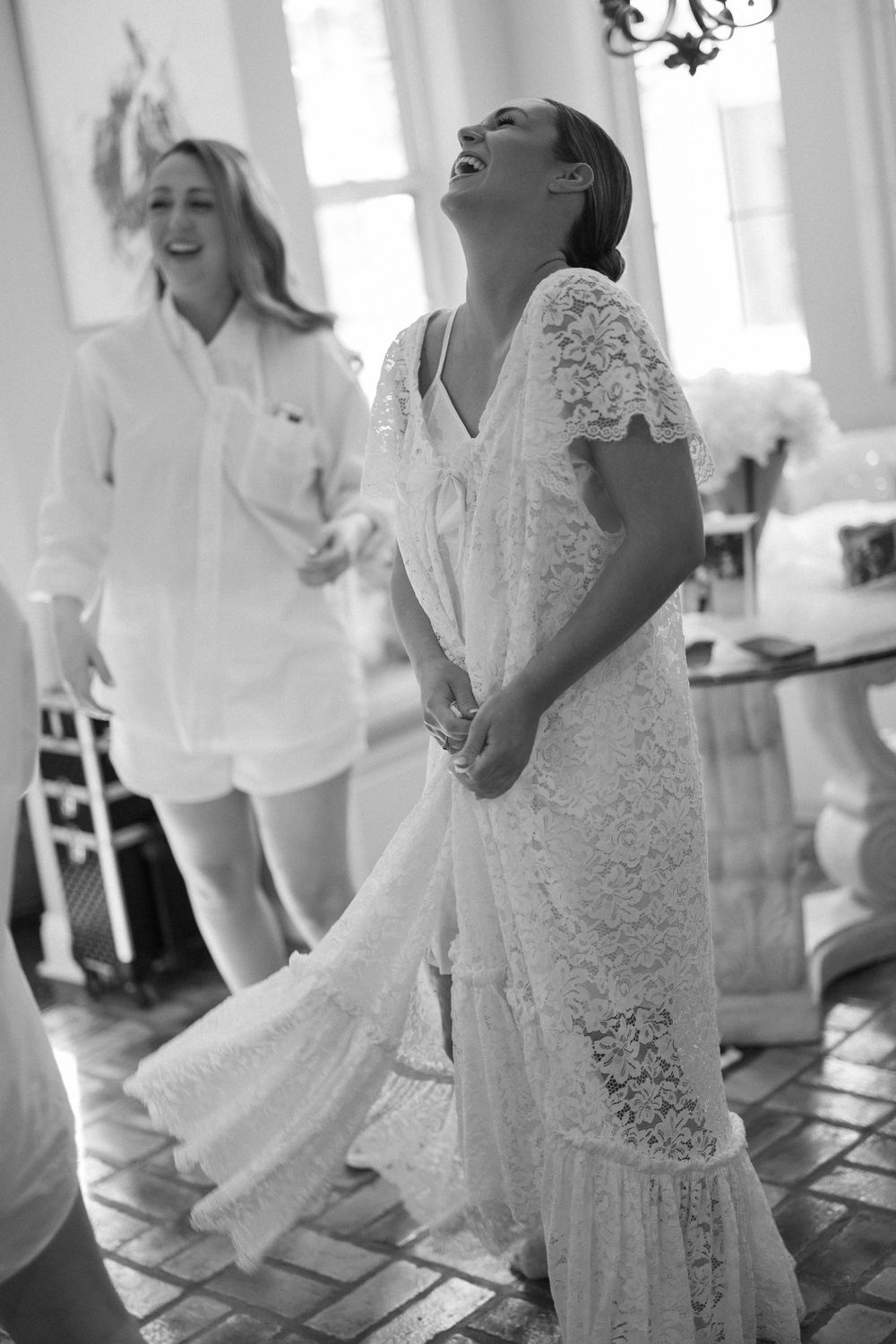 Black and white photo of bride in lace dress laughing joyfully during wedding preparations.