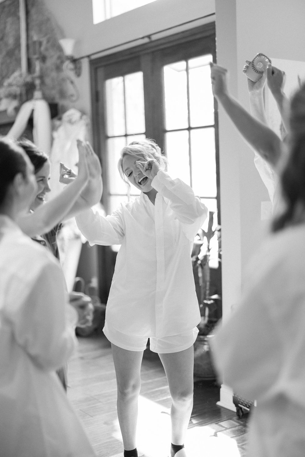 Black and white photo of bride in white shirt set celebrating with bridesmaids indoors.