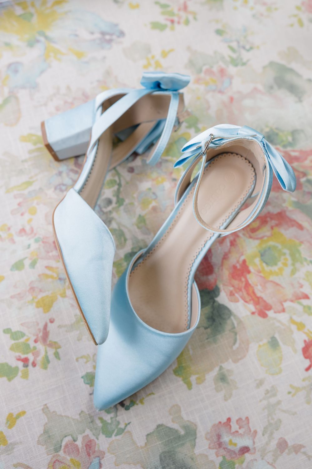 Light blue satin block heel wedding shoes with bow ankle straps on floral background.