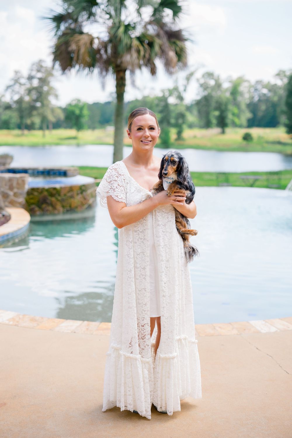 Smiling bride in white lace dress holding a dachshund dog near a pool outdoors.