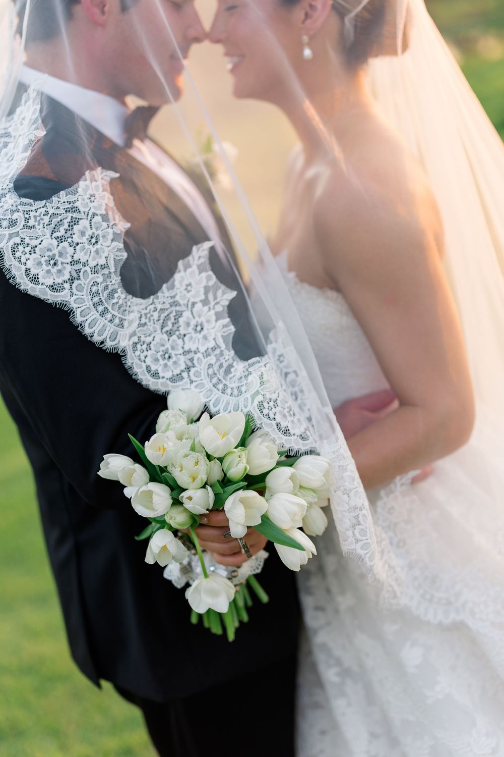 Bride and groom sharing an intimate moment under lace veil, bride holding white tulip bouquet in golden light.