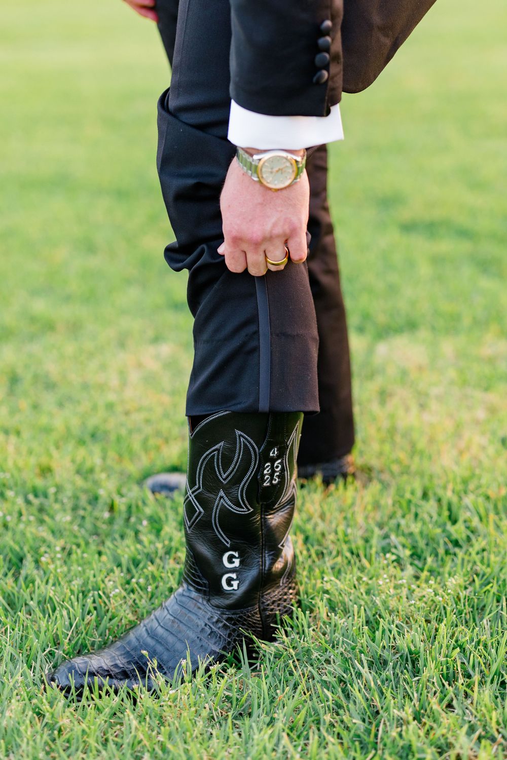 Groom wearing monogrammed black cowboy boots with gold watch and black suit standing on green grass.