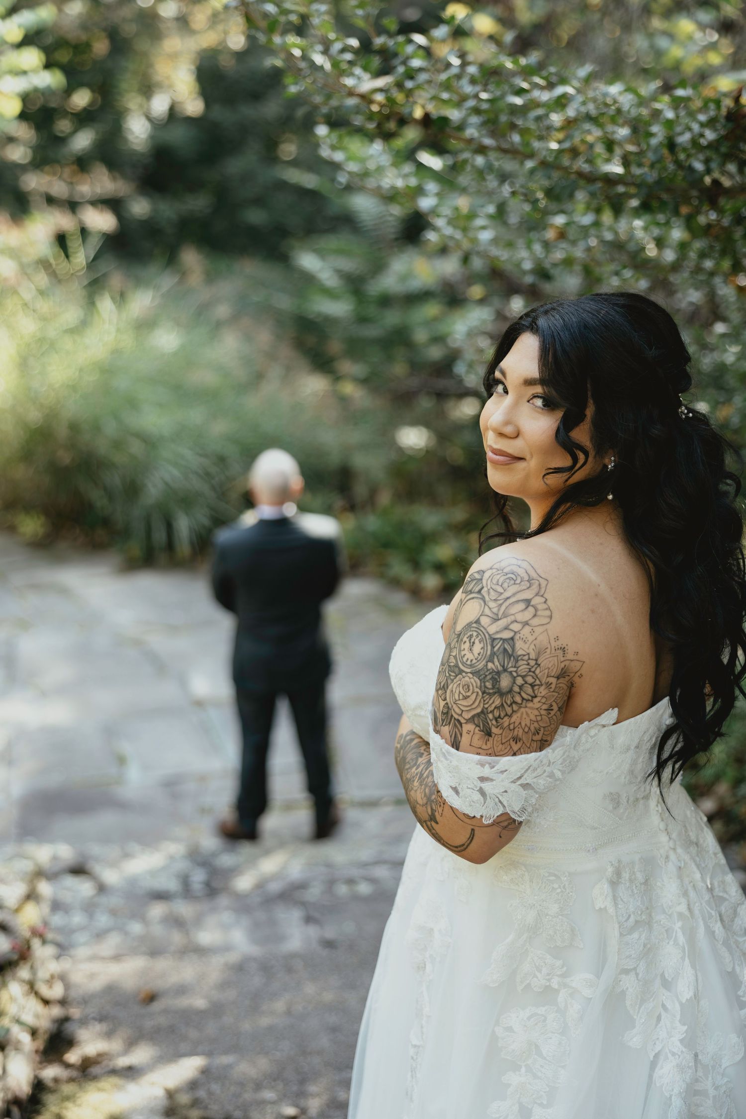 A wedding couple poses in a wooded outdoor setting with the bride wearing a white lace dress with visible tattoos.
