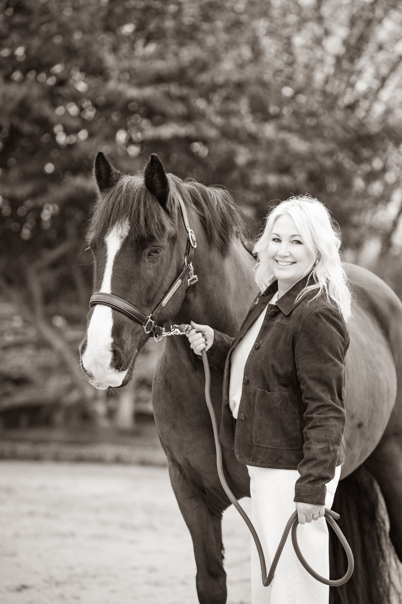 A beautiful bay horse wearing a black bridle stands next to someone in a white lace dress in an outdoor setting.