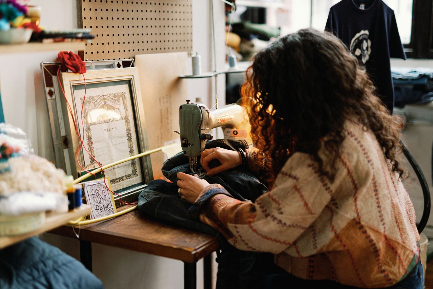 A person with curly hair works at a sewing machine in a crafting workspace with pegboard walls.