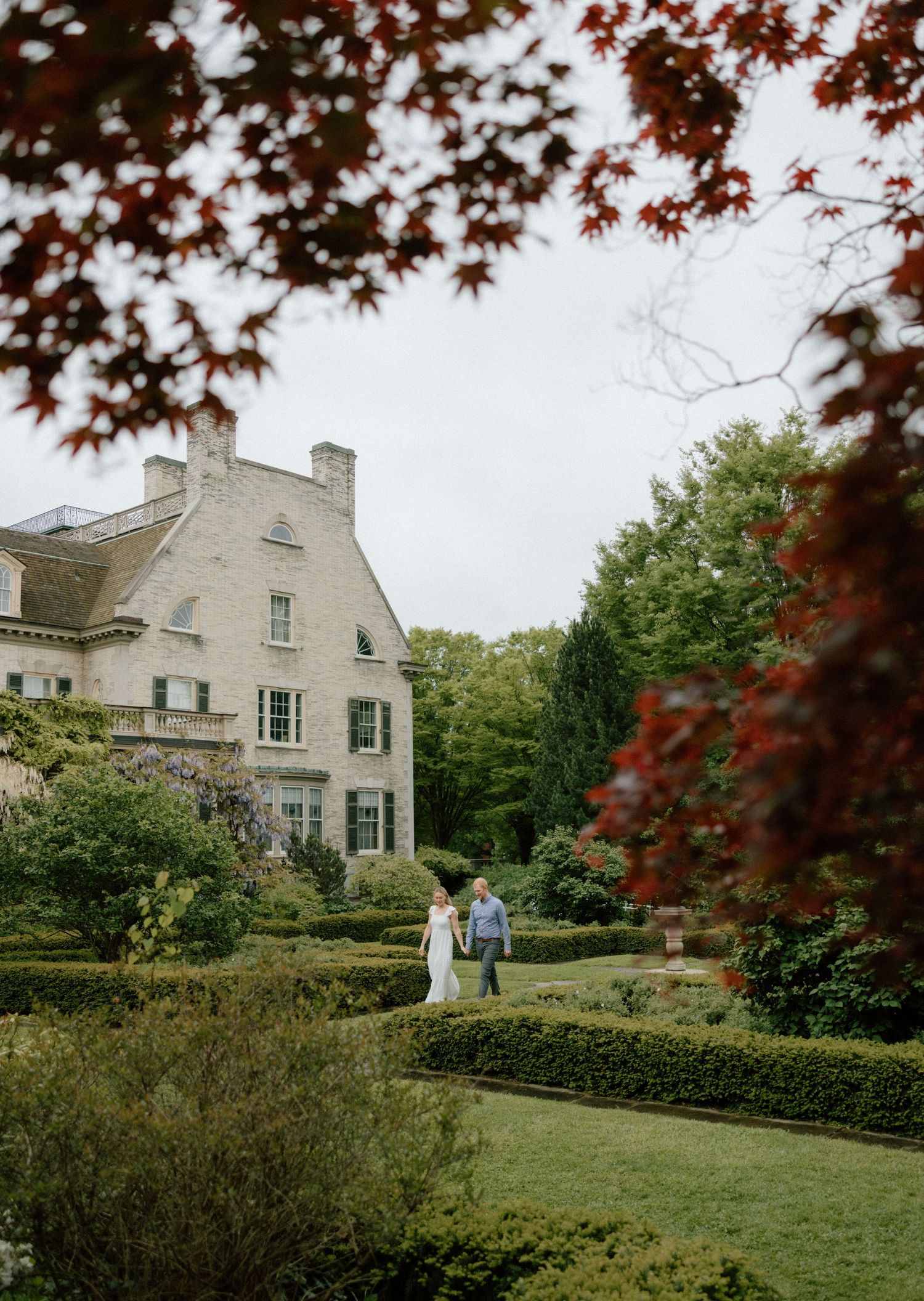 Elegant white mansion with manicured hedges and gardens framed by red maple leaves on a cloudy autumn day.