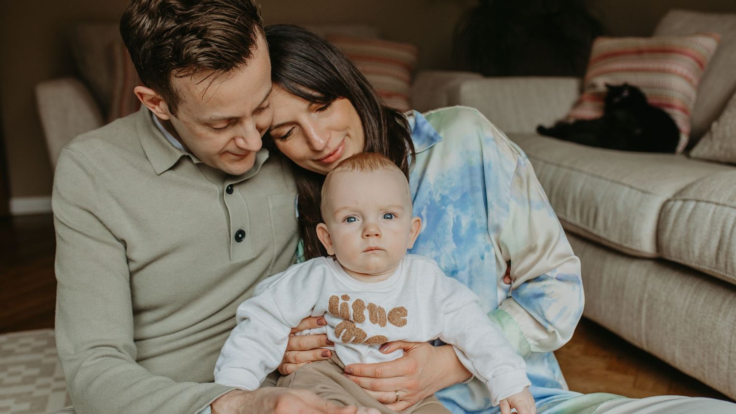 A couple snuggles with their baby on the floor at home while wearing casual clothing.