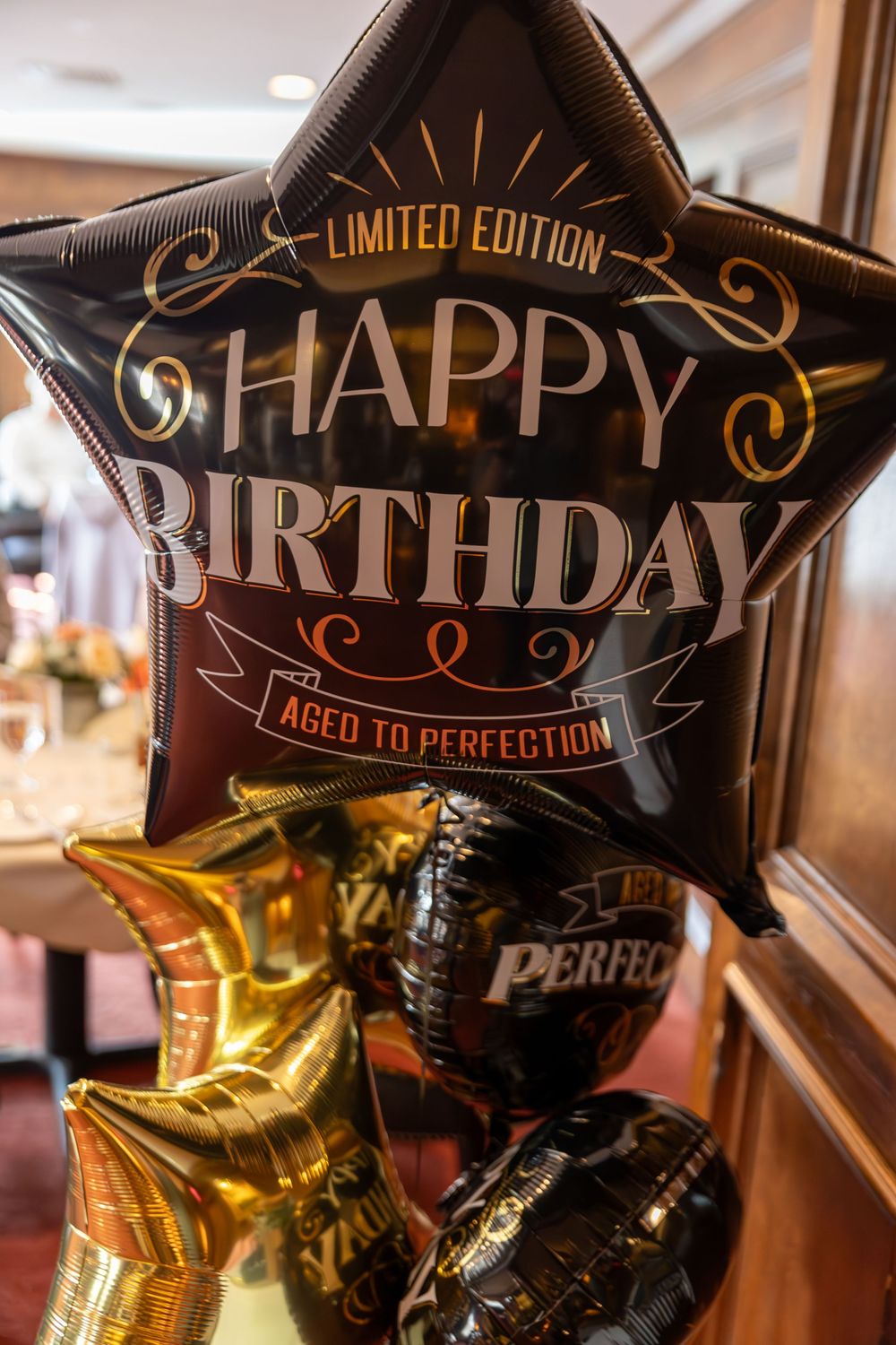 Black and gold birthday balloons with 'Aged to Perfection' text float against a warm interior background.