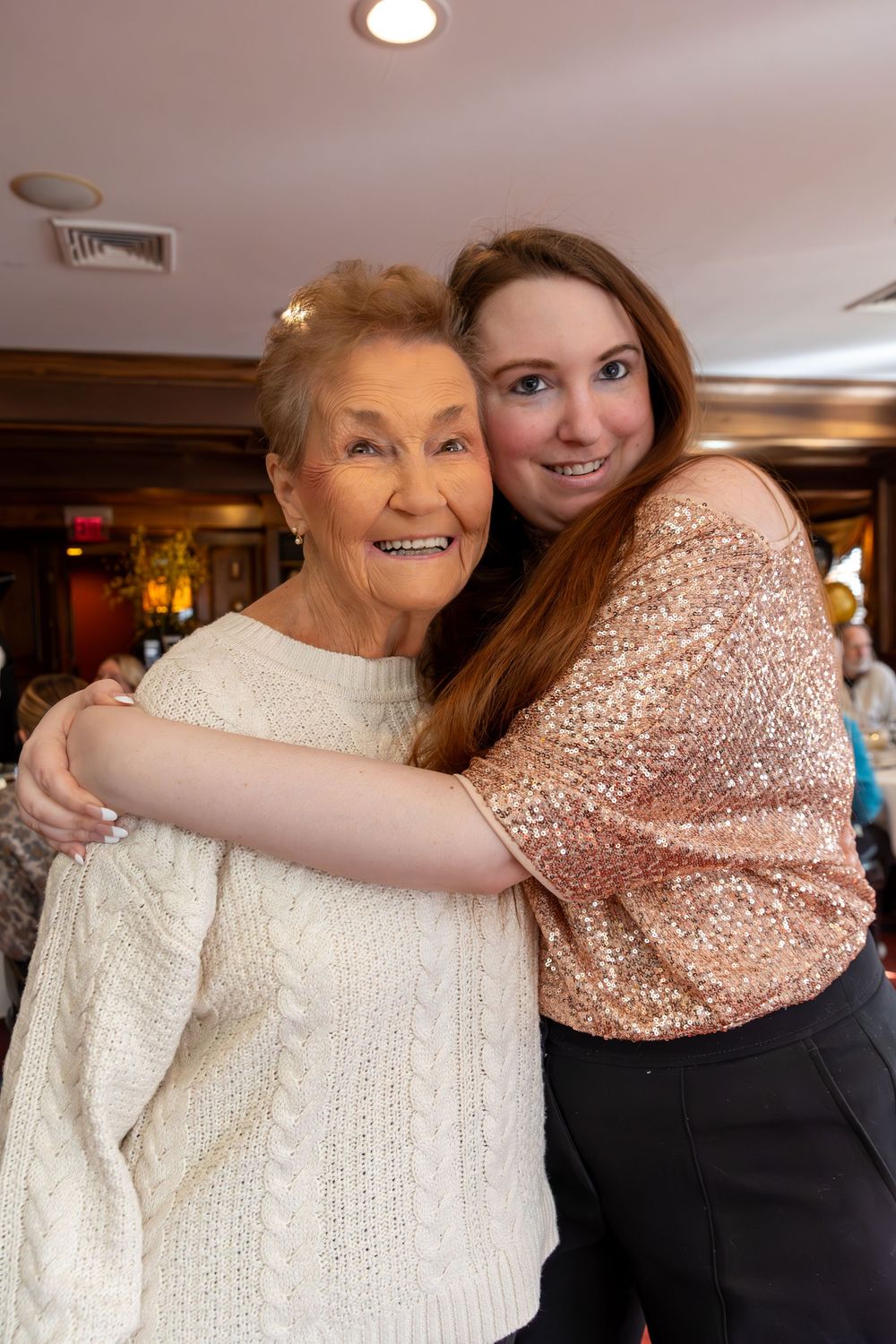 A heartwarming embrace between two people wearing a white knit sweater and a sparkly rose gold top at an indoor event.