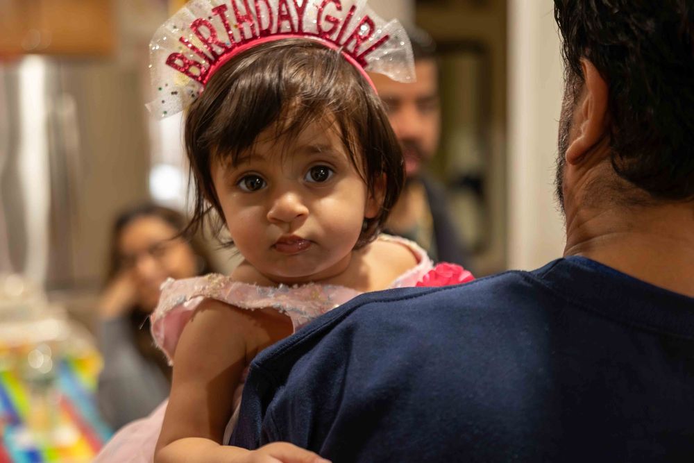A child wearing a birthday girl crown and pink dress celebrates at a party with colorful decorations in the background.