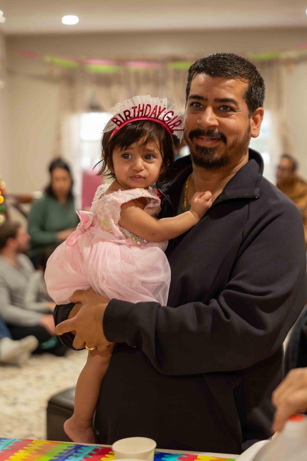 A young child in a pink dress and birthday crown is held at a birthday party celebration.