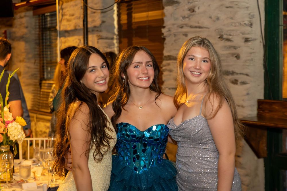 Three friends pose together at an elegant evening event with warm lighting and rustic stone walls in the background.