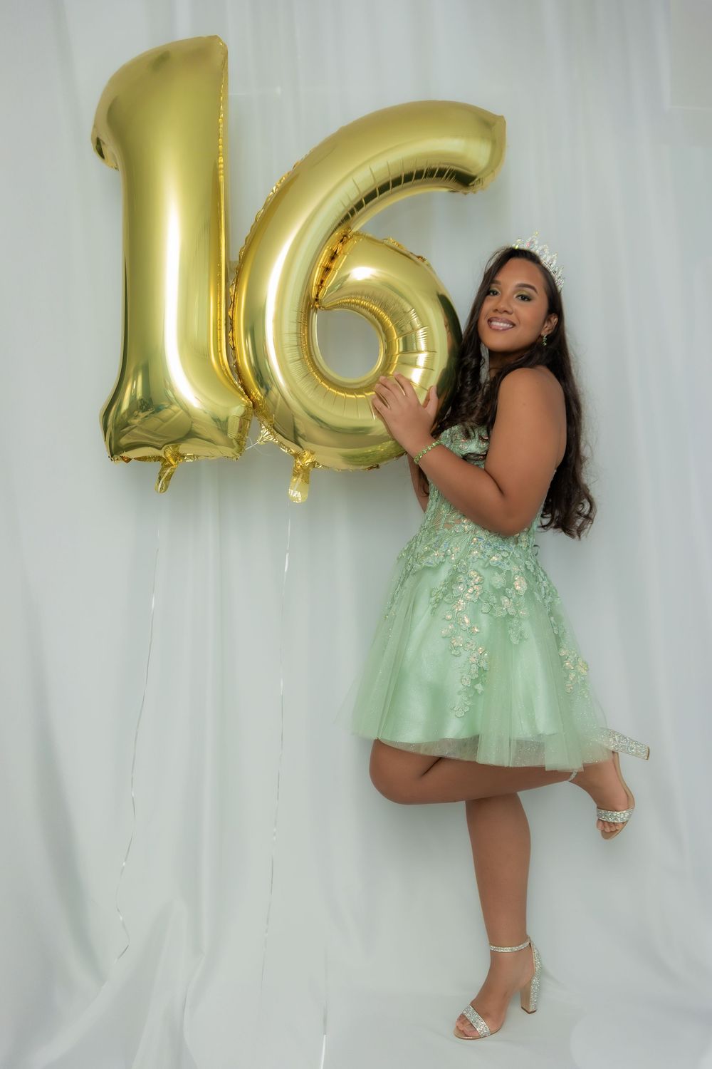 A person in a mint green dress poses playfully with large gold number balloons against a white backdrop.