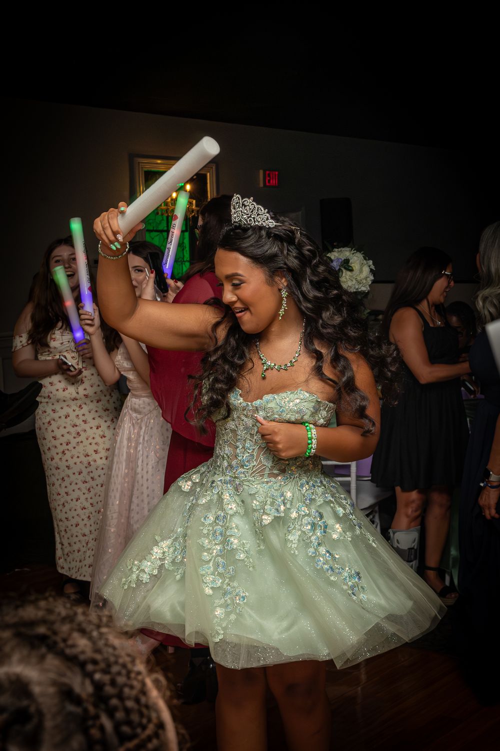 A dancer in a sparkly mint green dress twirls on a dark dance floor holding a glowing light stick.