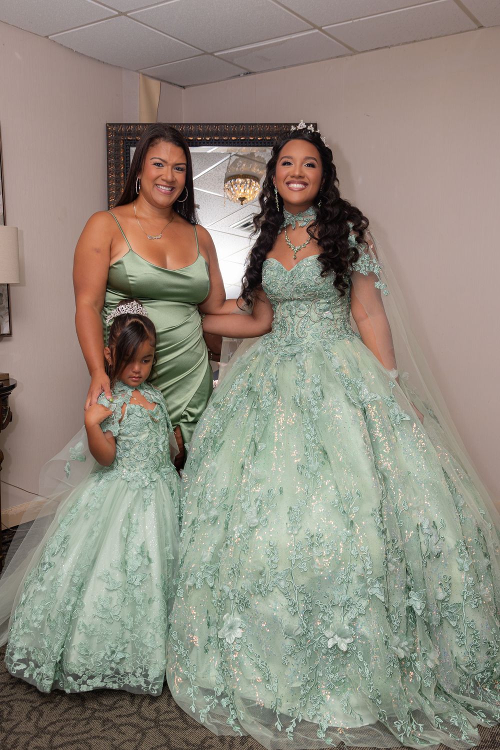 Three people wearing matching mint green dresses pose together in a room, with one in an elaborate ballgown with lace details.