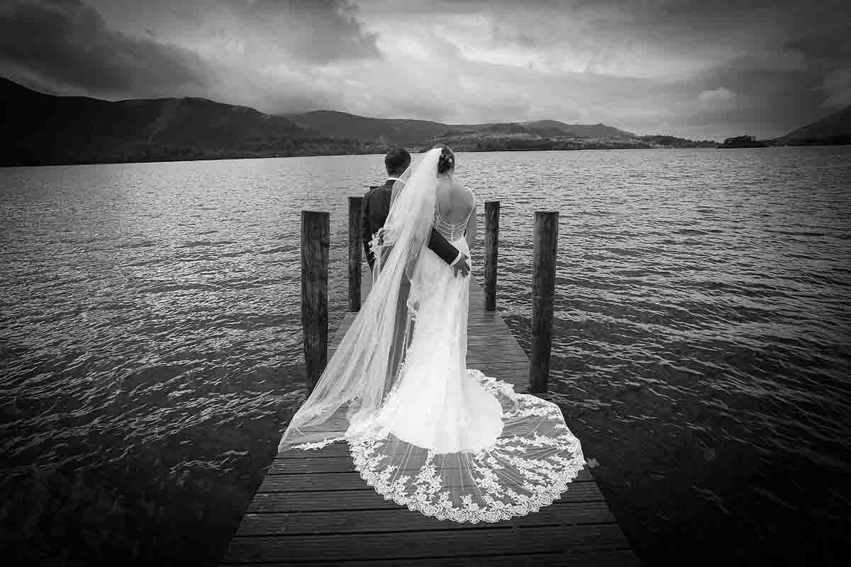 Black and white photo of a bride and groom standing on a wooden dock overlooking a serene lake on their wedding day.
