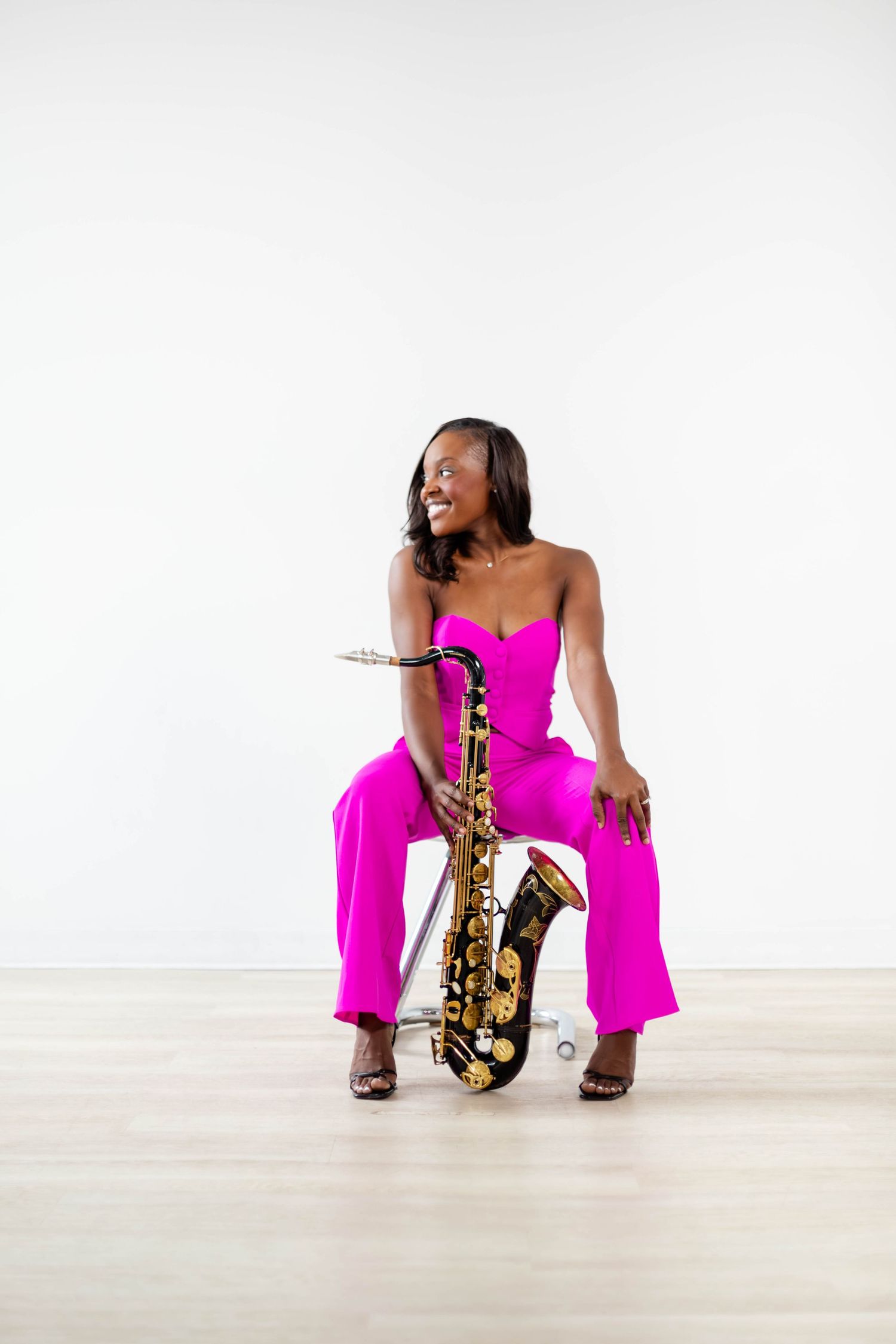 Musician in bright pink dress sits with saxophone in white studio setting creating artistic poses.