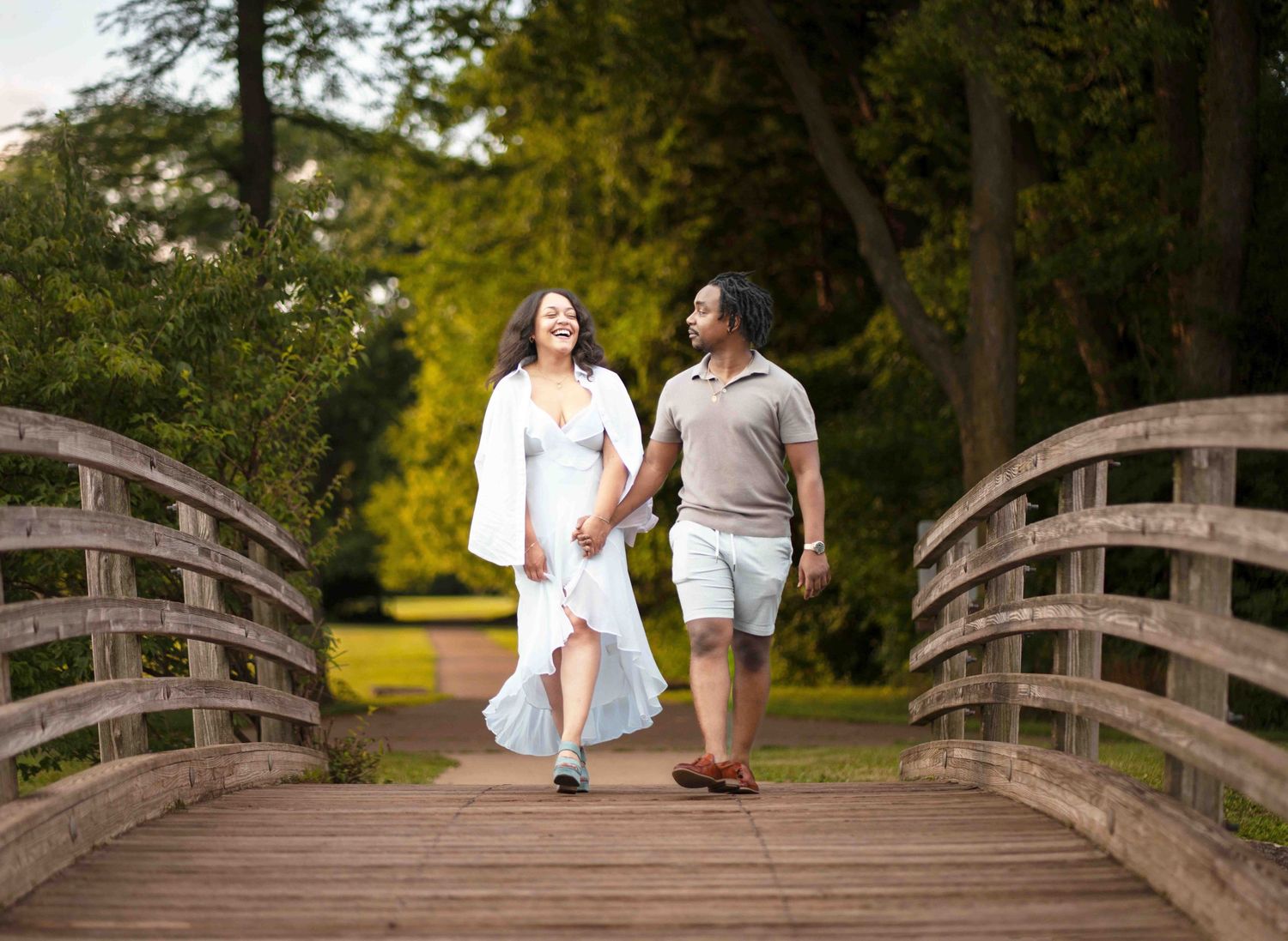 Couple walking together on a rustic wooden footbridge in a scenic park setting during golden hour.