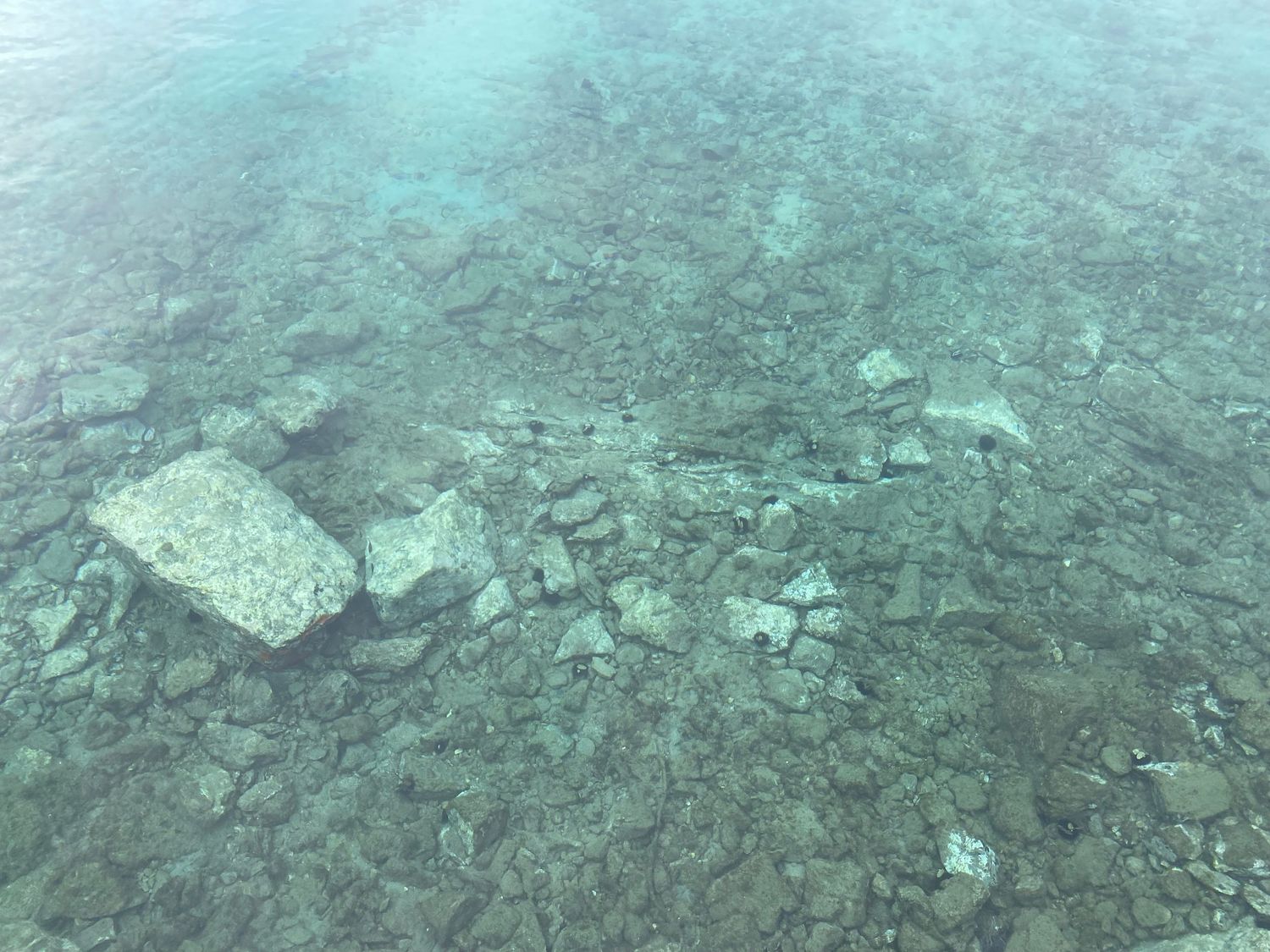 Underwater view of scattered rocks and debris on murky seafloor with blue-green tinted water.
