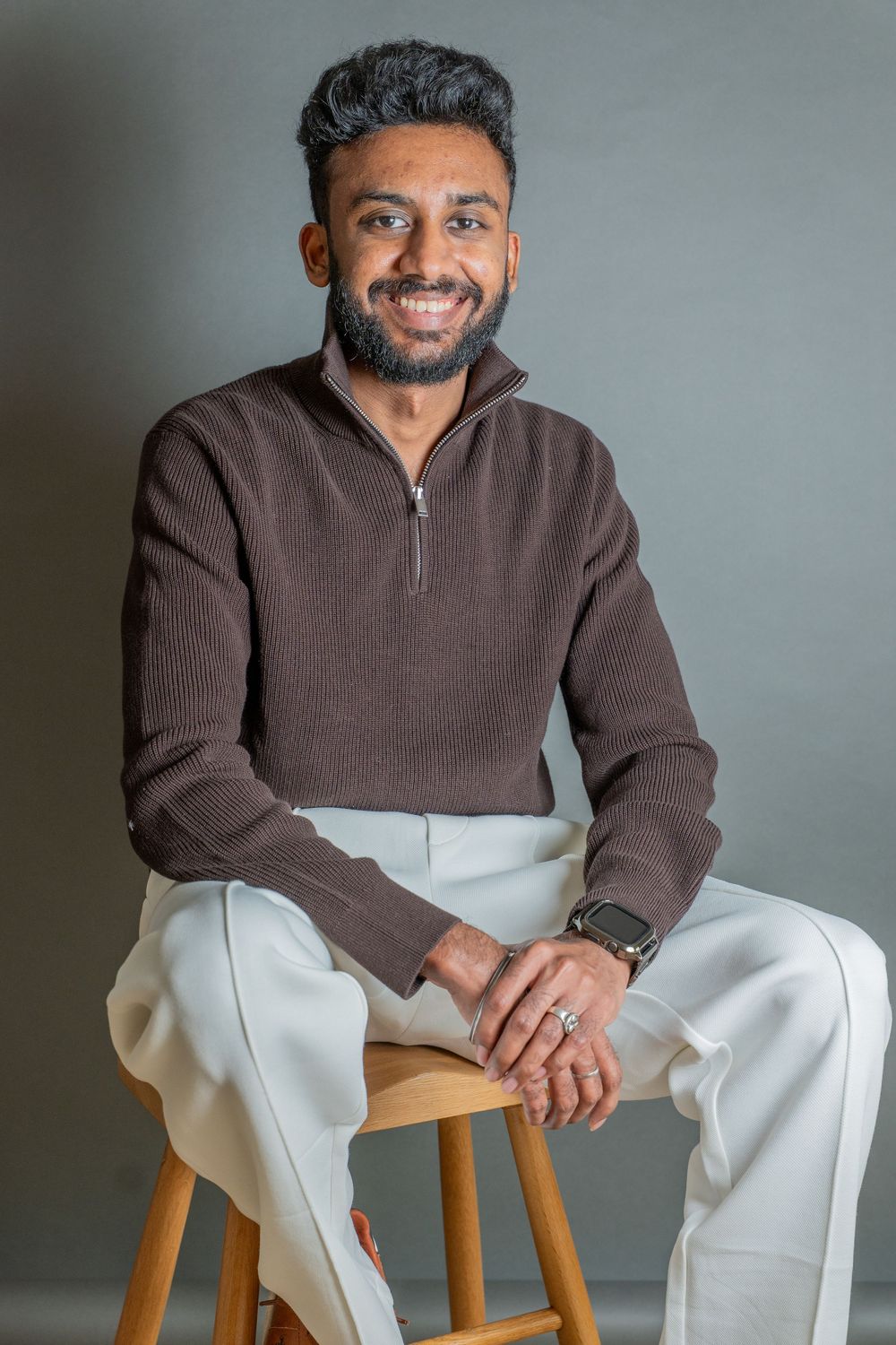 A person wearing a brown and white color block sweater poses on a wooden chair against a gray background.