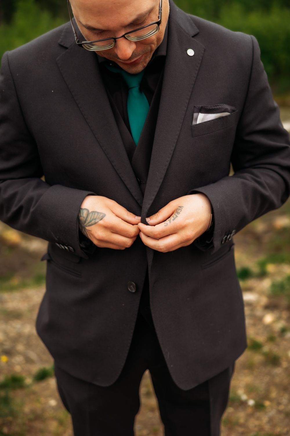 Groom in black suit with teal tie and tattooed hands buttons his jacket outdoors.