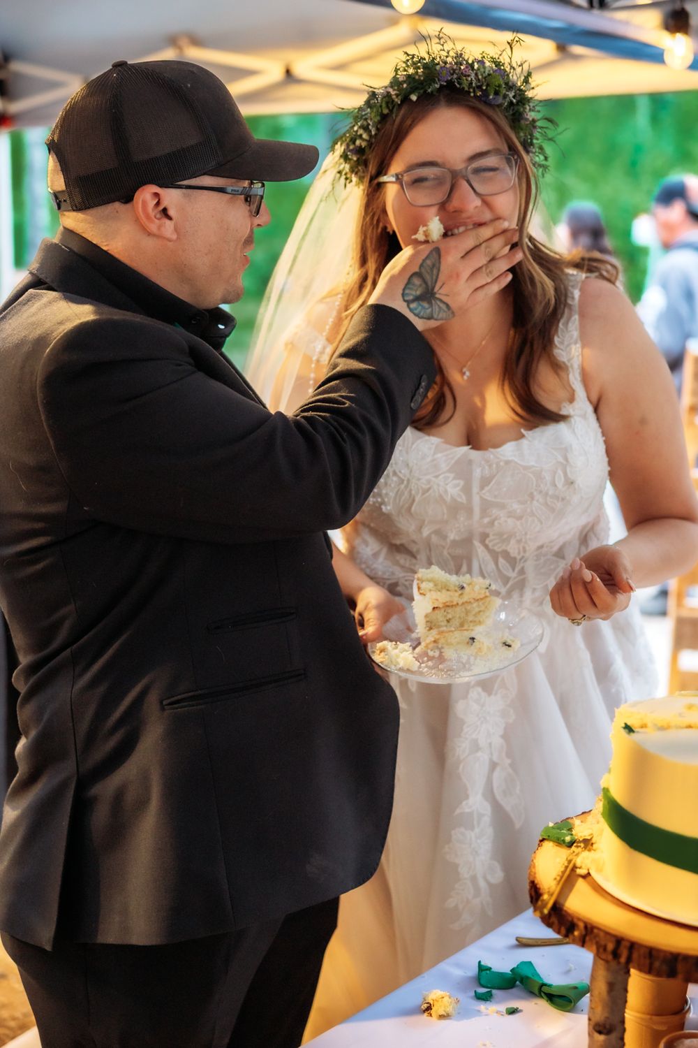 Groom feeds wedding cake to glasses-wearing bride in white dress and floral crown at outdoor reception.