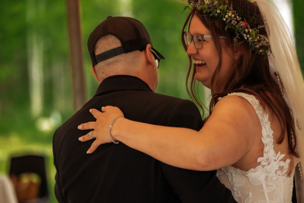 Bride in floral crown laughing joyfully while embracing groom in black shirt during outdoor wedding ceremony.